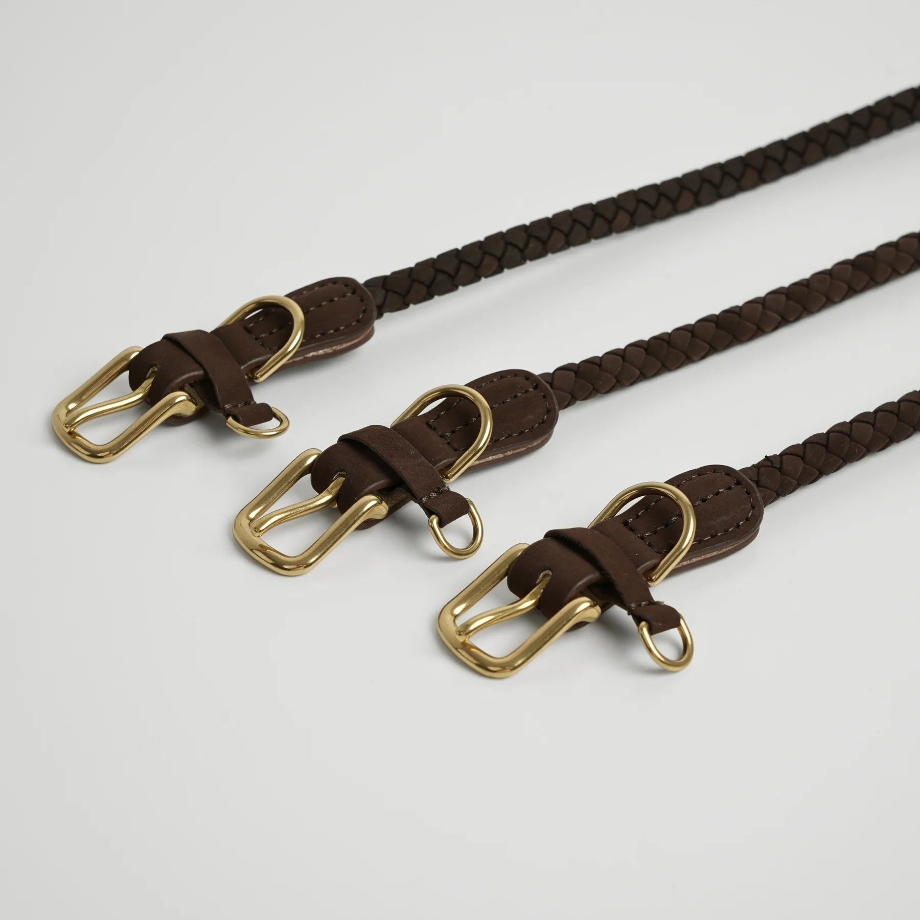 Three braided brown leather collars with gold buckles on a light gray background
